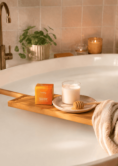 Luxuriöse Wellness-Badewanne mit milchigen Badewasser und der Leafy Milk & Honey Badewürfel-Verpackung auf einem Holztablett. Umgeben von Kerzenlicht und einem weichen Handtuch entsteht eine entspannende Spa-Atmosphäre.