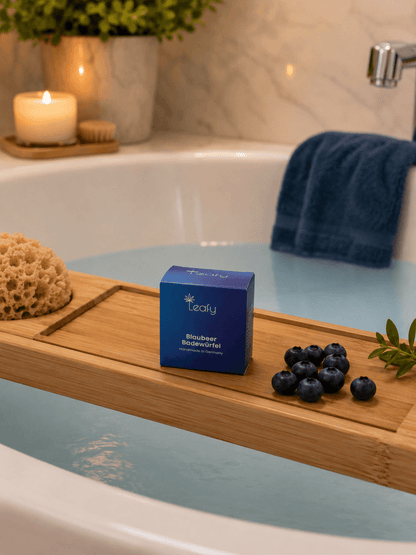 Modernes Lifestyle-Foto einer freistehenden weißen Badewanne mit milchig hellblauem Badewasser. Auf einem Bambus-Badewannenbrett steht die blaue Verpackung des Leafy Blaubeer Badewürfels vor einer brennenden Kerze und einem gefalteten Handtuch. Frische Blaubeeren sind dekorativ auf dem Brett arrangiert, jedoch nicht im Wasser.